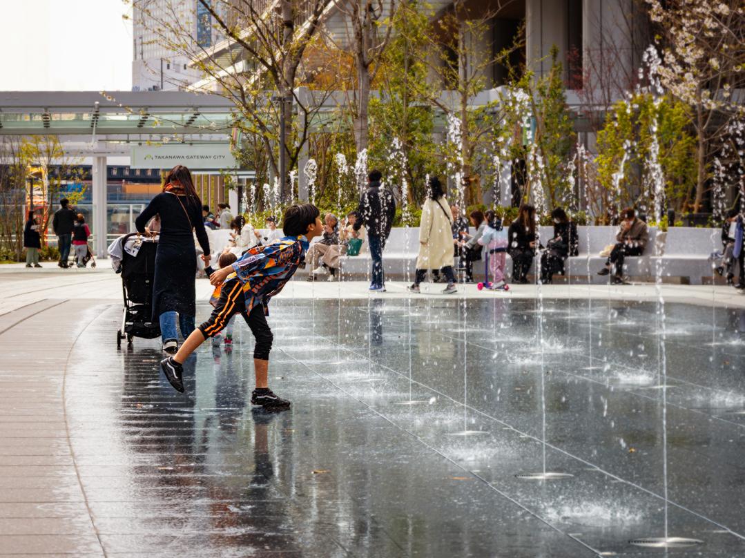Children playing in the water fountain feature in the public courtyard of Takanawa Gateway City.