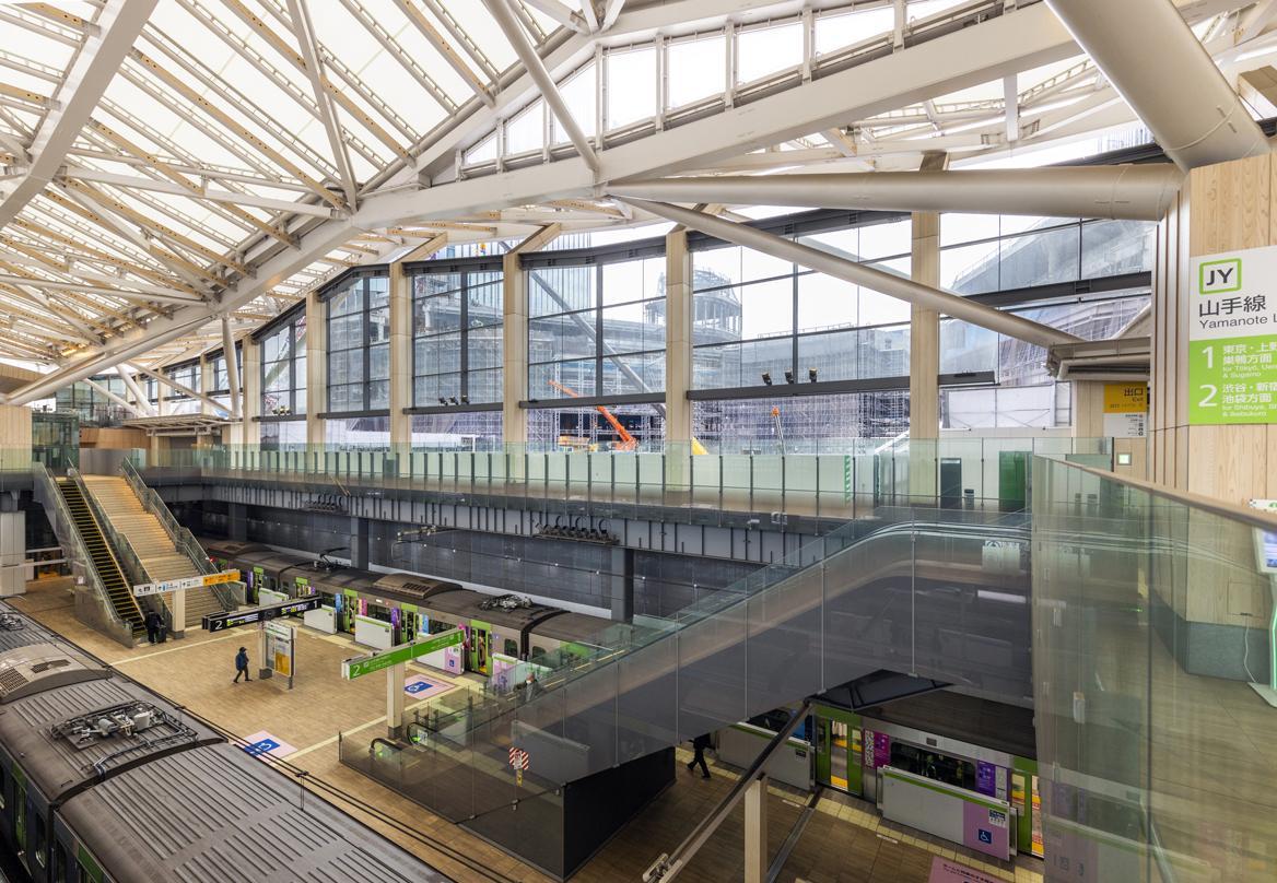 Interior view of the Takanawa Gateway City station under construction, featuring modern architecture with large glass windows, escalators, and platforms.