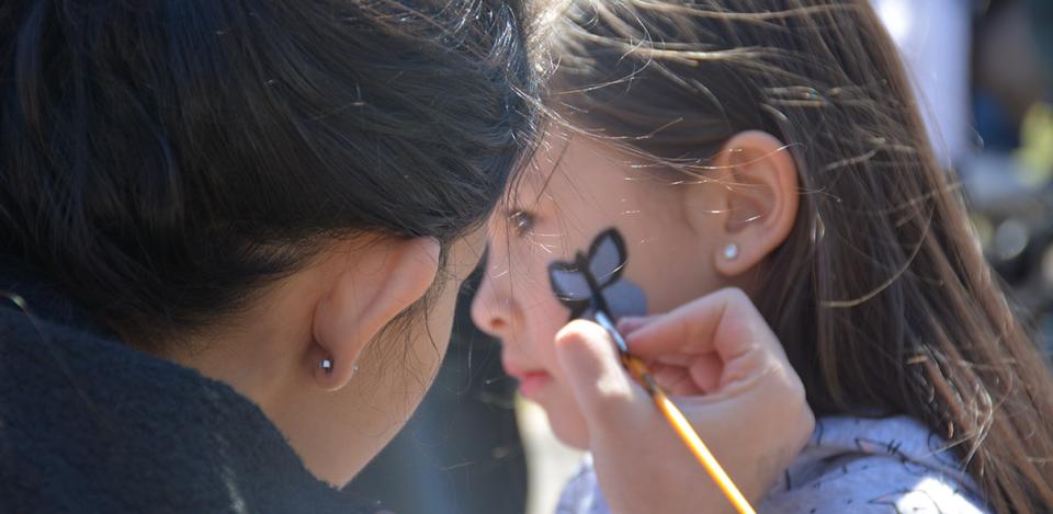 Woman face-painting a blue colored butterfly on a Asian woman’s face.