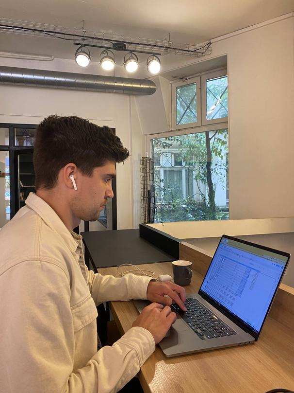 Owen focused on his laptop in a well-lit coworking space, earphones in, with a view of greenery through the window beside him