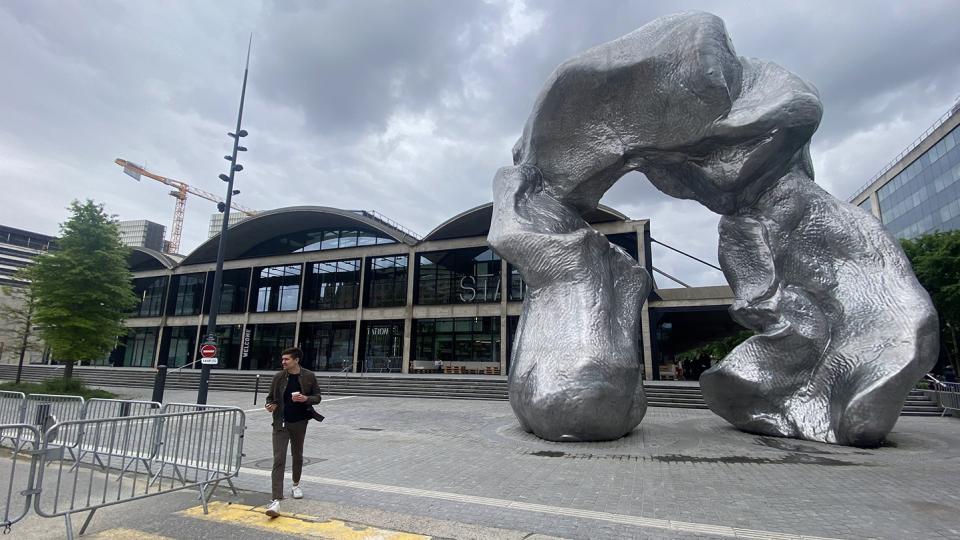 Owen walks past a striking, large-scale abstract sculpture in a London public square, with contemporary buildings and ongoing construction visible in the backdrop